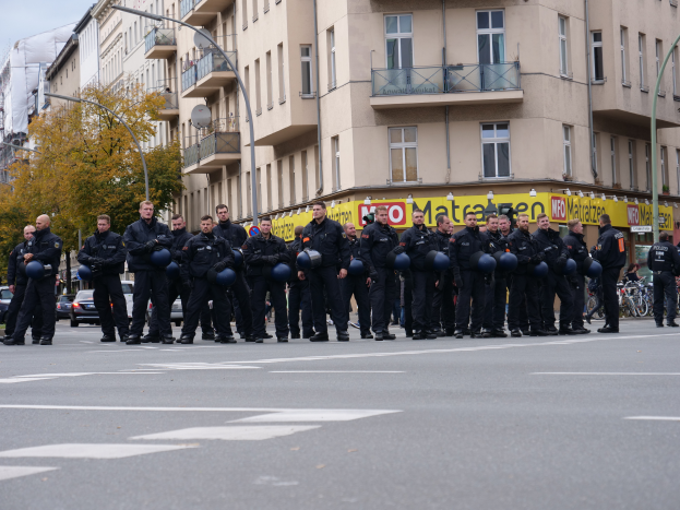 Eine Gruppe von Polizisten in schwarzen Uniformen mit blauen Helmen steht in der Mitte einer Straße, die von Laternenpfählen, Bäumen und Gebäuden mit Glasfenstern und Balkonen gesäumt ist, mit mehr Bäumen und einem klaren blauen Himmel im Hintergrund.