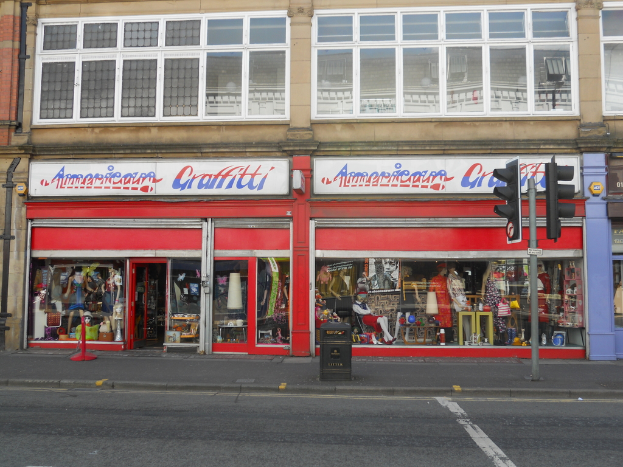 Eine Straßenszene mit einem Gebäude, mehreren Schaufensterpuppen, einem Müllcontainer, einem Pfahl, Verkehrsampeln und einer Sonnenbrille auf dem Boden.