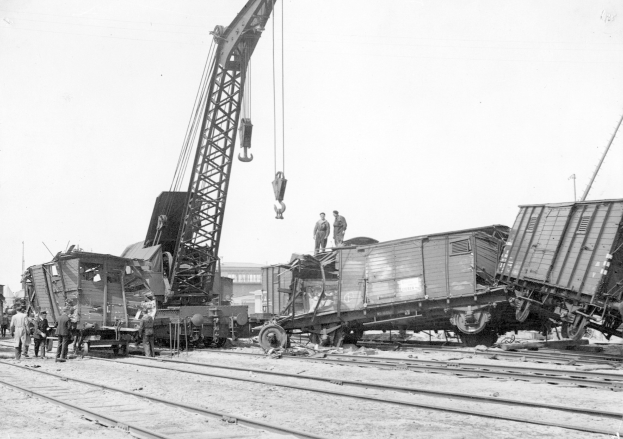 Schwarß-weißes Bild eines Zugunglücks mit einem Kran, der ein Waggon auf die Schienen hievt, mehreren Menschen in der Nähe und im Hintergrund Gebäude sowie einen klaren Himmel.