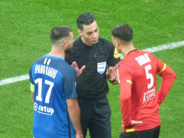 Schiedsrichter in schwarzer Uniform, der mit zwei Männern auf einem Fußballfeld spricht, einer in einem blauen und roten T-Shirt mit Text und Brille, der andere mit Brille.