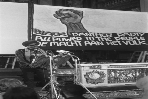 Ein Mann in einer schwarzen Jacke steht auf einer Bühne und hält ein Mikrofon, mit einem "Black Panther Party"-Plakat dahinter und mehreren Mikrofonständern drumherum, auf einem Schwarz-Weiß-Bild.