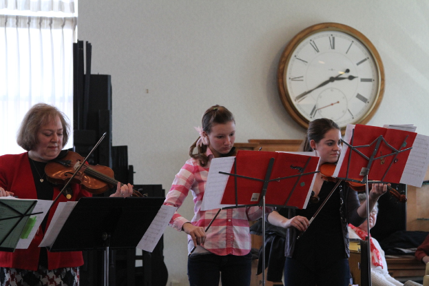 Drei Frauen auf der Bühne mit Geigen spielend, vor sich Notenständer und Noten; im Vordergrund sitzt eine Frau und hört zu; an der weißen Wand hängt eine Uhr, und im linken Eck ist ein weißer Vorhang zu sehen.