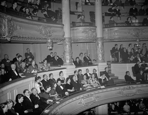 Ein Schwarz-Weiß-Foto einer großen Menschenmenge in einem Theater, mit einigen Stehenden und einigen auf der Treppe Sitzenden, Blumensträußen, Geländern, Säulen und Kronleuchtern im Hintergrund und Text unten.