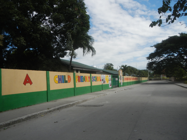 Eine Straße mit einer grünen und gelben Wand, die ein "Kindergartenfreundliche Schule"-Schild zeigt, umgeben von Bäumen, Häusern, Polen und einem bewölkten Himmel.