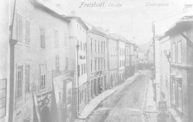 Ein altes Schwarz-Weiß-Foto einer Straße in Freistadt, mit Gebäuden auf beiden Seiten der Straße und einem klaren Himmel im Hintergrund, mit Text über dem Bild.