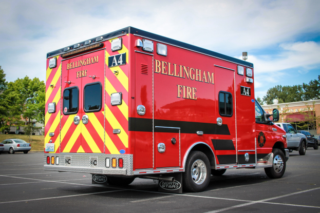 Ein Rettungswagen des Bellingham Fire Department, der auf einem Parkplatz mit Bäumen und Gebäuden geparkt ist, unter einem klaren blauen Himmel.