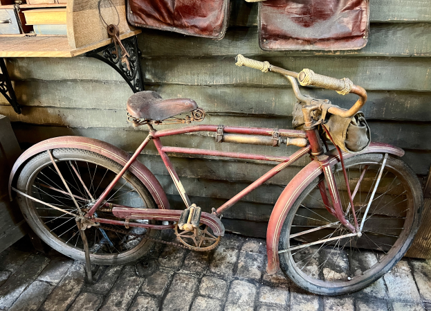 Ein altes rotes Fahrrad in schlechtem Zustand, das Rost und Verfärbungen zeigt, steht vor einer hölzernen Wand. Links davon steht ein Tisch mit ein paar Gegenständen, und rechts hängen mehrere Taschen an der Wand.