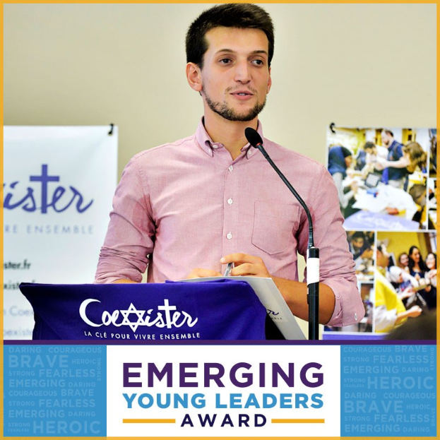 Ein Mann steht an einem Rednerpult mit einem Mikrofon, hält ein Blatt Papier in der Hand und steht vor einer Banner mit der Aufschrift "Emerging Young Leaders Award", während mehrere Personen rechts von ihm zu sehen sind.