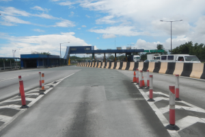 Eine Straße mit Verkehrskegeln am Rand, eine Mittelinsel, Fahrzeuge, Laternenpfähle, Bäume, eine Brücke und ein bewölkter Himmel im Hintergrund.