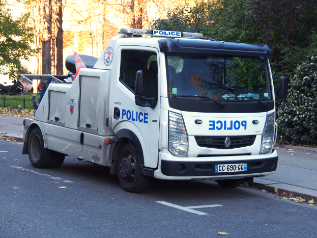 Ein Polizeilaster steht am Straßenrand geparkt, umgeben von Bäumen, Pflanzen, Gras, einem Zaun und einem Gebäude im Hintergrund.