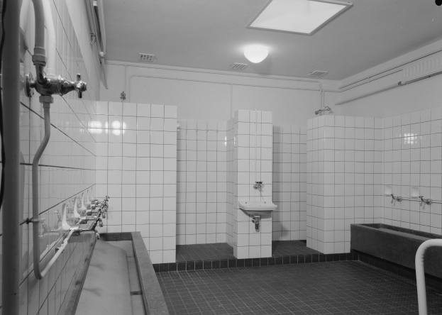 Ein Schwarz-Weiß-Foto einer öffentlichen Toilette mit gefliesten Wänden, ausgestattet mit Waschbecken, einer Badewanne, freiliegenden Rohren und einer Deckenlampe.