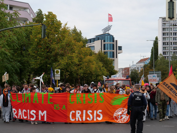 Eine Gruppe von Menschen hält ein Transparent mit der Aufschrift "Klimakrise ist eine Krise" beim Spaziergang auf einer baumbestandenen Straße mit parkenden Fahrzeugen, Schildern und Gebäuden unter einem klaren blauen Himmel.