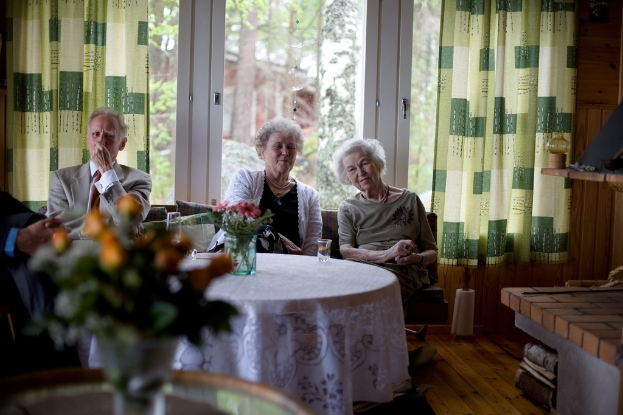Eine Gruppe älterer Personen auf einem Sofa mit einem Tisch davor, der eine Vase und ein Glas hat, und eine Glastür mit Vorhängen dahinter.