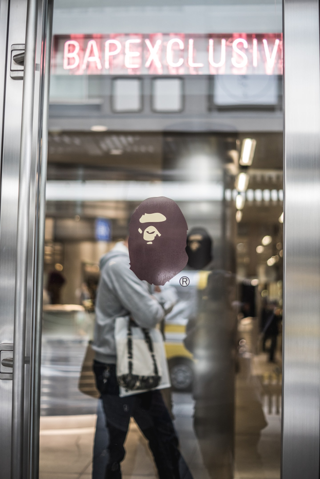 Eine Person mit Maske steht vor einer Glastür mit der Aufschrift "Bape Exclusive" und einem beleuchteten Gebäude im Hintergrund.