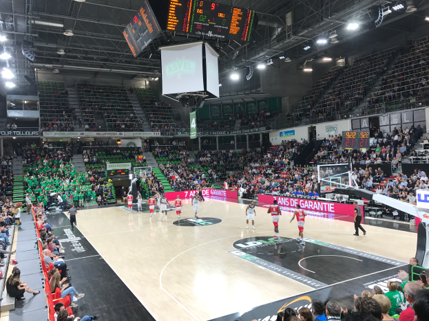 Ein Basketball-Spiel in einer großen Arena mit Zuschauern auf den Rängen, Anzeigetafeln mit "Asvel vs Galatasaray" und einer Decke mit Lampen und einem Bildschirm.