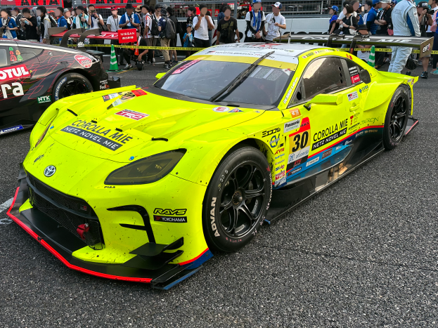 Ein Toyota Supra GT3 Rennwagen auf der Straße, umgeben von einer Menschenmenge, einige machen Fotos, mit Absperrungen, Schildern und Verkehrskegeln im Hintergrund unter einem klaren blauen Himmel.