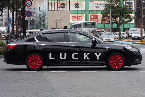 Ein schwarzes Auto mit dem Wort "Lucky" darauf fährt eine Straße entlang, umgeben von anderen Fahrzeugen, Bäumen, Polen, Schildern, Pflanzen und Gebäuden.