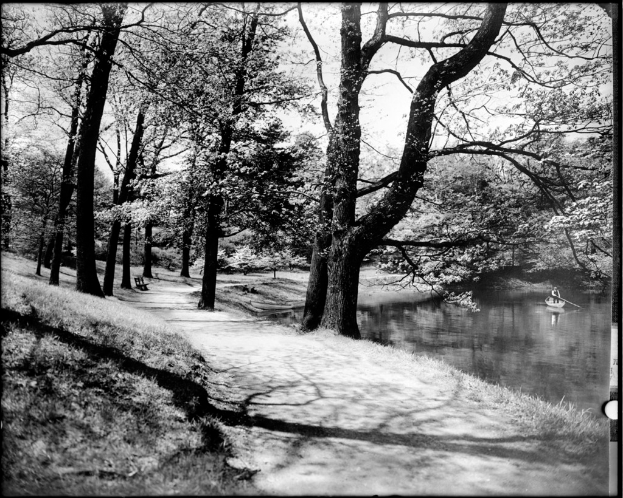 Ein Schwarz-Weiß-Bild, das eine Person zeigt, die ein Boot auf dem Wasser rudert, mit Bäumen, Gras, einer Bank und Himmel im Hintergrund.