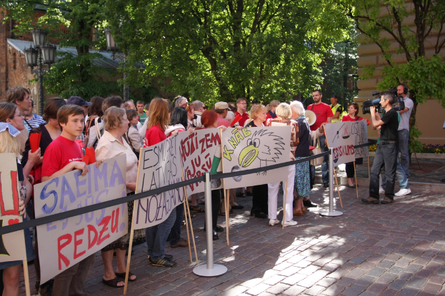 Eine Gruppe von Menschen mit Schildern auf einem Gehweg, mit Männern, die Kameras halten, Laternen, Bäumen und Gebäuden im Hintergrund.