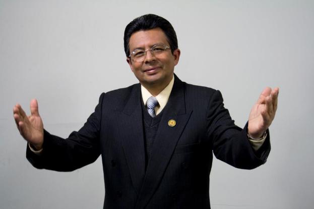 Ein Mann im Anzug mit Krawatte und ausgebreiteten Armen, der eine Brille trägt, steht vor einem weißen Hintergrund und sieht ernst aus.