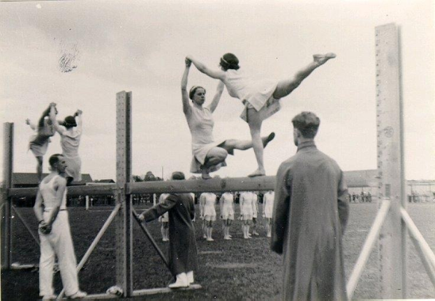 Schwarz-weißes Foto von Turnern auf einem Balancebalken mit Zuschauern und einer Hintergrundlandschaft aus Gebäuden, Bäumen und Himmel.