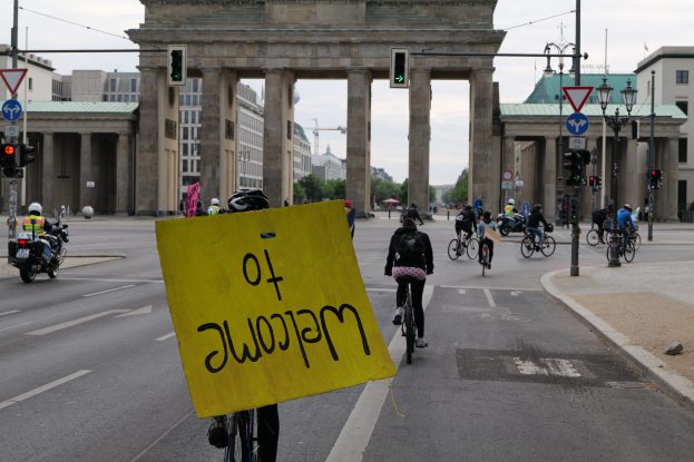 Eine Gruppe von Menschen auf Fahrrädern fährt eine Straße vor dem Brandenburger Tor in Berlin, Deutschland, mit Helmen, wobei eine Person ein gelbes Schild hält, umgeben von Laternenmasten, Verkehrsampeln, Gebäuden, Bäumen und einem klaren blauen Himmel.