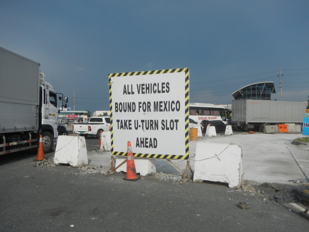 Lkw auf einem Parkplatz neben einem Schild "Alle Fahrzeuge nach Mexiko" geparkt mit verstreuten Verkehrskegeln und Schildern, Gebäuden und Bäumen im Hintergrund unter einem klaren blauen Himmel.