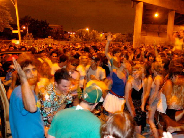 Leute tanzen im Vordergrund bei einem Konzert mit Lichtern, Pfählen, Bäumen und anderen Gegenständen im Hintergrund.