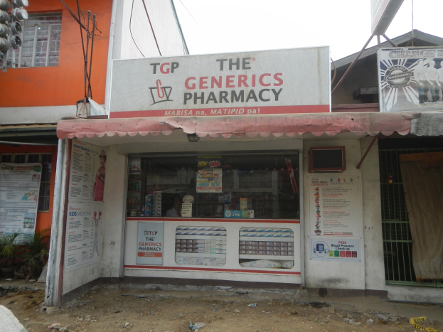 Außenansicht der Generics Pharmacy Filiale mit umliegenden städtischen Elementen unter einem klaren blauen Himmel.