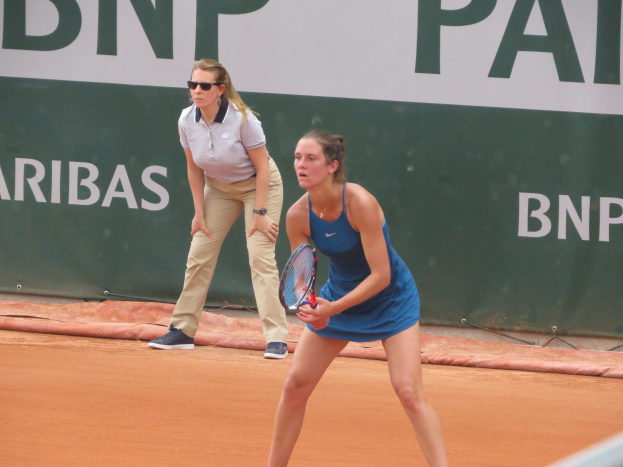 Zwei Frauen beim Tennis auf einem Sandplatz, wobei eine in einem blauen Kleid mit einem Schläger in der Hand im Vordergrund steht und eine andere hinter ihr steht, während ein grüner Banner mit Text im Hintergrund sichtbar ist.