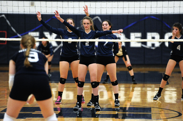 Drei Mädchen auf einem Volleyballfeld mit erhobenen Händen, ein Netz davor und ein Zaun im Hintergrund.