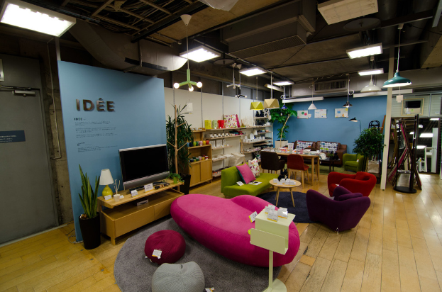 Ein Schaufenster mit Haushaltsgeräten und Möbeln, darunter ein Fernseher auf einem Tisch, eine rosafarbene Couch, ein Sofa, zwei Stühle um einen kleinen Hocker, ein Esstisch mit Stühlen, ein großes Sofa im Hintergrund und verschiedene Artikel auf Regalen, alles von Deckenleuchten beleuchtet.