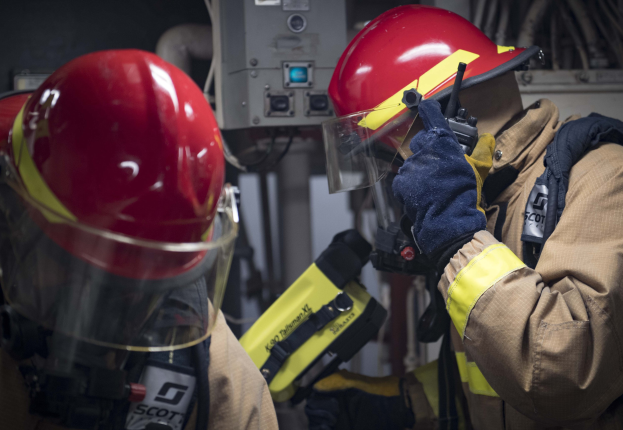 Zwei Feuerwehrleute in Schutzausrütstüzen arbeiten an einem Feuerhydranten während einer Ausbildung mit Maschinen und Kabeln im Hintergrund.