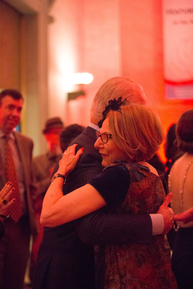 Zwei Menschen umarmen sich herzlich auf einer Party, umgeben von anderen, mit einer beleuchteten Wand im Hintergrund.