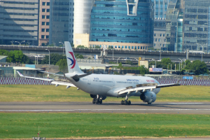 Großraumjet steigt von einer Flughafenpiste mit umgebenem Gras, Zaun, Bäumen, Straßenlaternen, Schildern, einer Brücke, Gebäuden mit Fenstern und einem bewölkten Himmel auf.