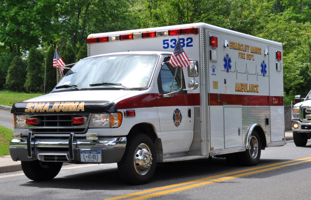 Ein Rettungswagen steht am Straßenrand mit zwei Flaggen im Hintergrund, umgeben von Bäumen unter einem klaren blauen Himmel.