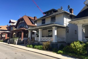 Eine Reihe von Höusern mit Fenstern, Sülen, Gelöndern, Stufen, Döchern, mit einem Straßlenmast, Pflanzen, einem Zaun, einem Weg, Böumen und einem bewölktem Himmel, mit einem Kran auf einem Dach, der auf Bauarbeiten hinweist.