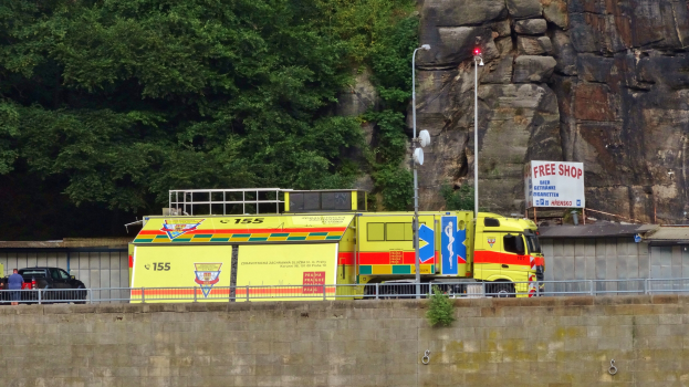 Ein Rettungswagen steht am Straßenrand neben einer Klippe, mit verschiedenen Gegenständen im Vordergrund und Bäumen und einem Auto im Hintergrund.