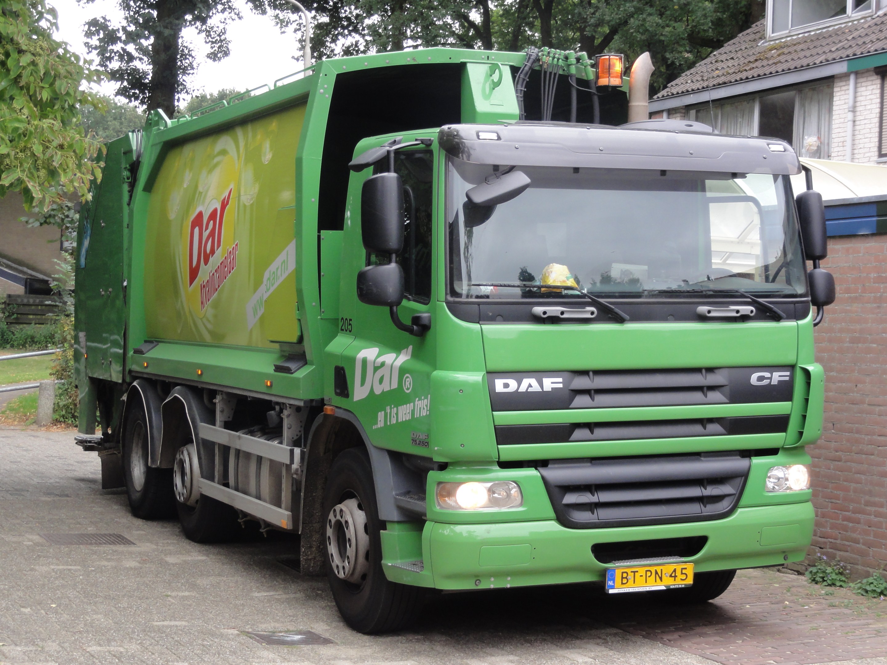 Ein grüner Müllwagen vor einem Backsteingebäude mit Bäumen, Pflanzen, Gras, einem Laternenmast und einem klaren blauen Himmel im Hintergrund.