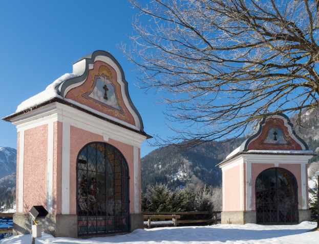 Eine kleine, schneebedeckte Kapelle mit Fenstern und T├╝ren, umgeben von einem Zaun und B├Ąumen, in einer verschneiten Wiese mit H├╝geln und einem bew├Âlktem Himmel im Hintergrund.