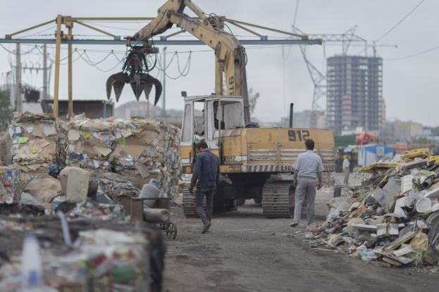 Eine Gruppe von Männern geht auf einer Schotterstraße neben einem Müllhaufen, mit einem Bagger im Hintergrund, Gebäude, Bäume und andere Gegenstände in der Umgebung.