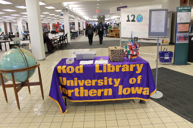 Eine Ausstellung mit einem Tisch, auf dem verschiedene Landesflaggen, ein Karton, Papiere und eine Fahne sowie links daneben eine Weltkugel, rechts ein Anschlagbrett mit einigen Menschen in der Nähe und viele Tische und Stühle im Hintergrund zu sehen sind.