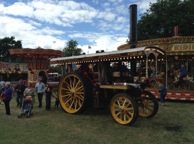 Eine Dampflokomotive steht auf einer grünen Fläche bei einem Volksfest, umgeben von Menschen und Kinderwagen, während im Hintergrund ein Karussell und Bäume unter einem bewölkten Himmel zu sehen sind.