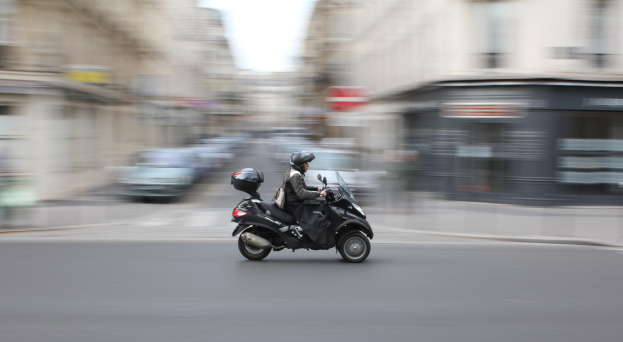 Eine Person in schwarzer Kleidung und Helm auf einem Motorroller auf einer Stadtstraße mit Fahrzeugen, Gebäuden und einem klaren blauen Himmel im Hintergrund.