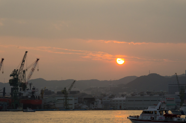 Mehrere Schiffe im Wasser, Kräne und Türme im Hintergrund, Gebäude, Berge, die Sonne und ein klarer Himmel.