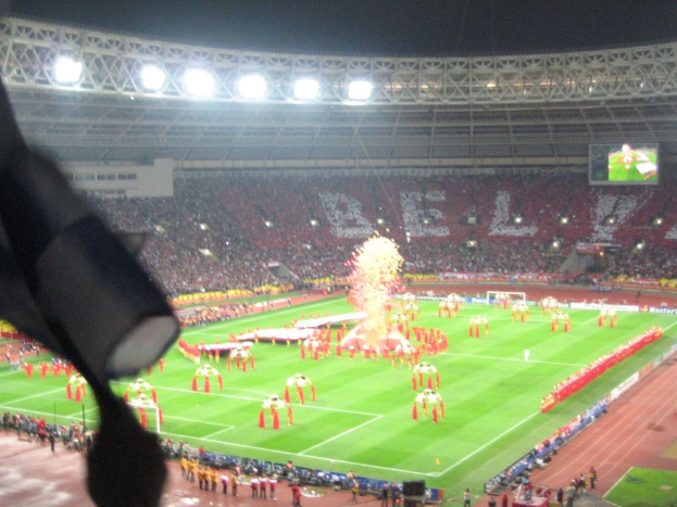 Ein Stadion mit Flutlicht, Eisengräben, Zuschauern, stehenden Personen, Werbetafeln, Anzeigetafeln, Überwachungskameras, Flaggen und Décorations.