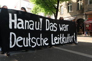 Eine Gruppe von Menschen geht auf einer Straße, die eine schwarze Fahne mit der Aufschrift "Hanau das war deutsche Leitkultur" trägt, mit Gebäuden, Bäumen und einem klaren blauen Himmel im Hintergrund.