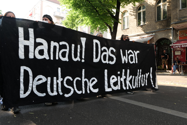 Eine Gruppe von Menschen geht auf einer Straße, die eine schwarze Fahne mit der Aufschrift "Hanau das war deutsche Leitkultur" trägt, mit Gebäuden, Bäumen und einem klaren blauen Himmel im Hintergrund.