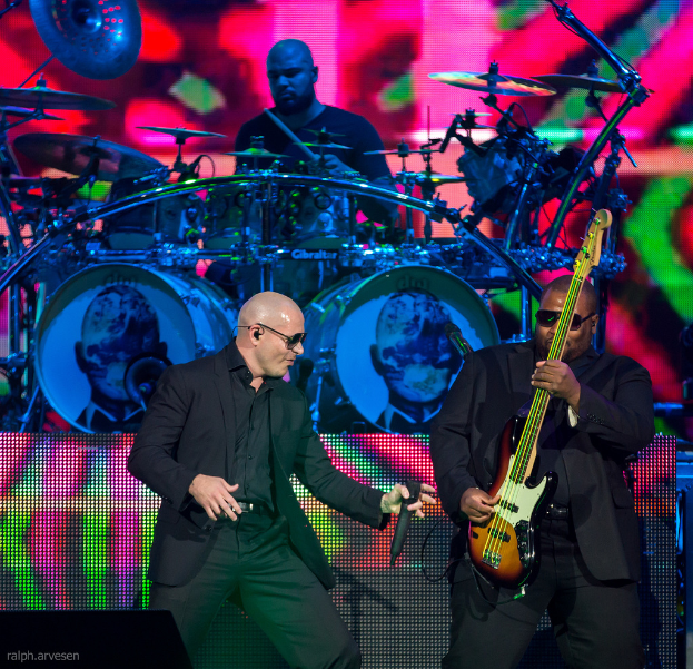 Ein Mann in der rechten Ecke spielt eine Gitarre, ein anderer Mann in einem schwarzen Blazer hält ein Mikrofon und lächelt, und ein Mann in einem schwarzen T-Shirt spielt Schlagzeug, alles vor einem bunten Hintergrund bei einem Musikkonzert.