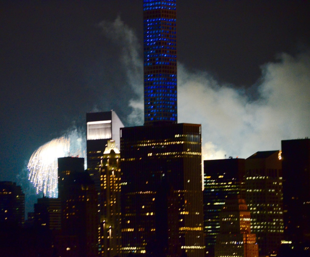 Geb√§ude mit Lichtern im Vordergrund, Feuerwerk und Rauch im Hintergrund, unter einem Himmel.